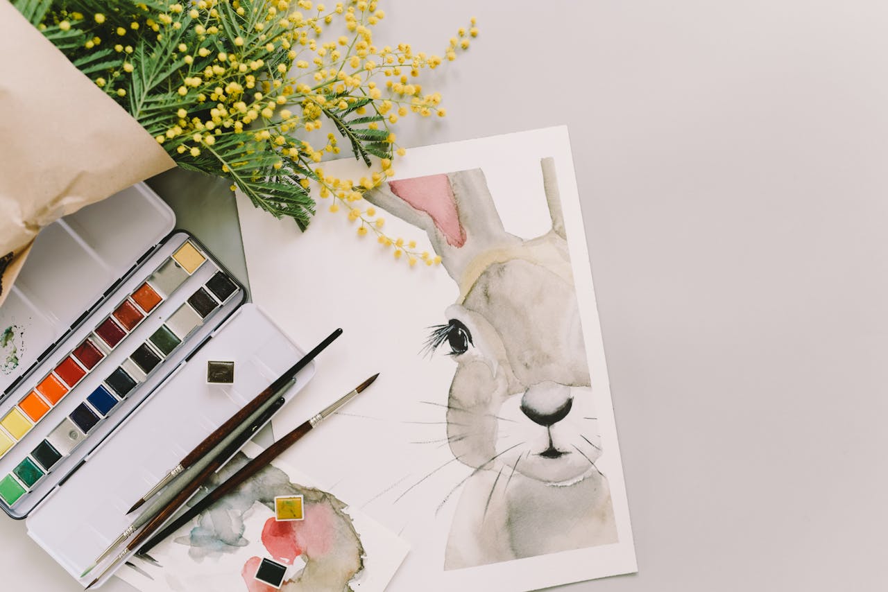 Home Watercolor painting of a bunny surrounded by mimosa flowers and art supplies.